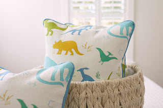 Decorative pillows from the Kids Collection: Dinos Pillow are charmingly displayed in a woven basket, featuring delightful dinosaur designs in blue, yellow, and green on a crisp white background. A window with shutters softly blurs into the backdrop, enhancing the rooms cozy charm.