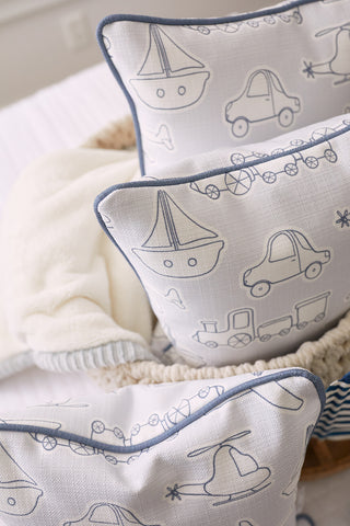 The Kids Collection: Transportation Pillow features three white pillows with blue outlines adorned with car, boat, train, and helicopter designs. They sit on a cozy bed with a soft white blanket, creating a playful and comfortable atmosphere.