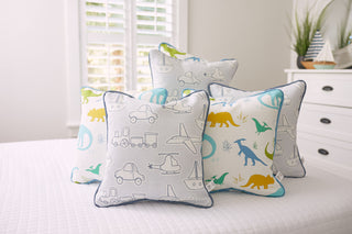 The Kids Collection: Transportation Pillow set includes five neatly arranged pillows with vehicle, dinosaur, and nautical designs. They rest on a white bed, surrounded by white furniture and a shuttered window.