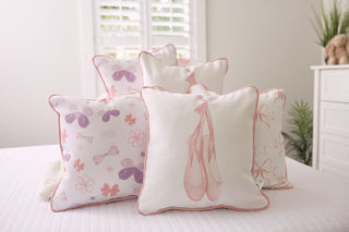 A neatly arranged set of Kids Collection: Butterfly Pillows, featuring ballet-themed designs with pink and purple ballet shoes and floral patterns, adorns a bed with white sheets in a soft, neutral-toned nursery accented by a shuttered window and plants.