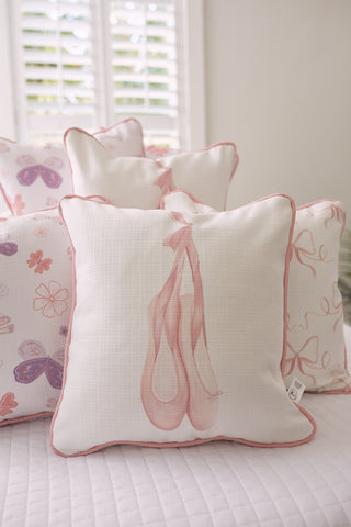 A Kids Collection: Bow & Ballet Pillow, featuring pink ballet slipper prints, is surrounded by floral and butterfly pillows. Soft light streams through window blinds in the background, creating a serene ambiance akin to a Sewing Down South® pattern.