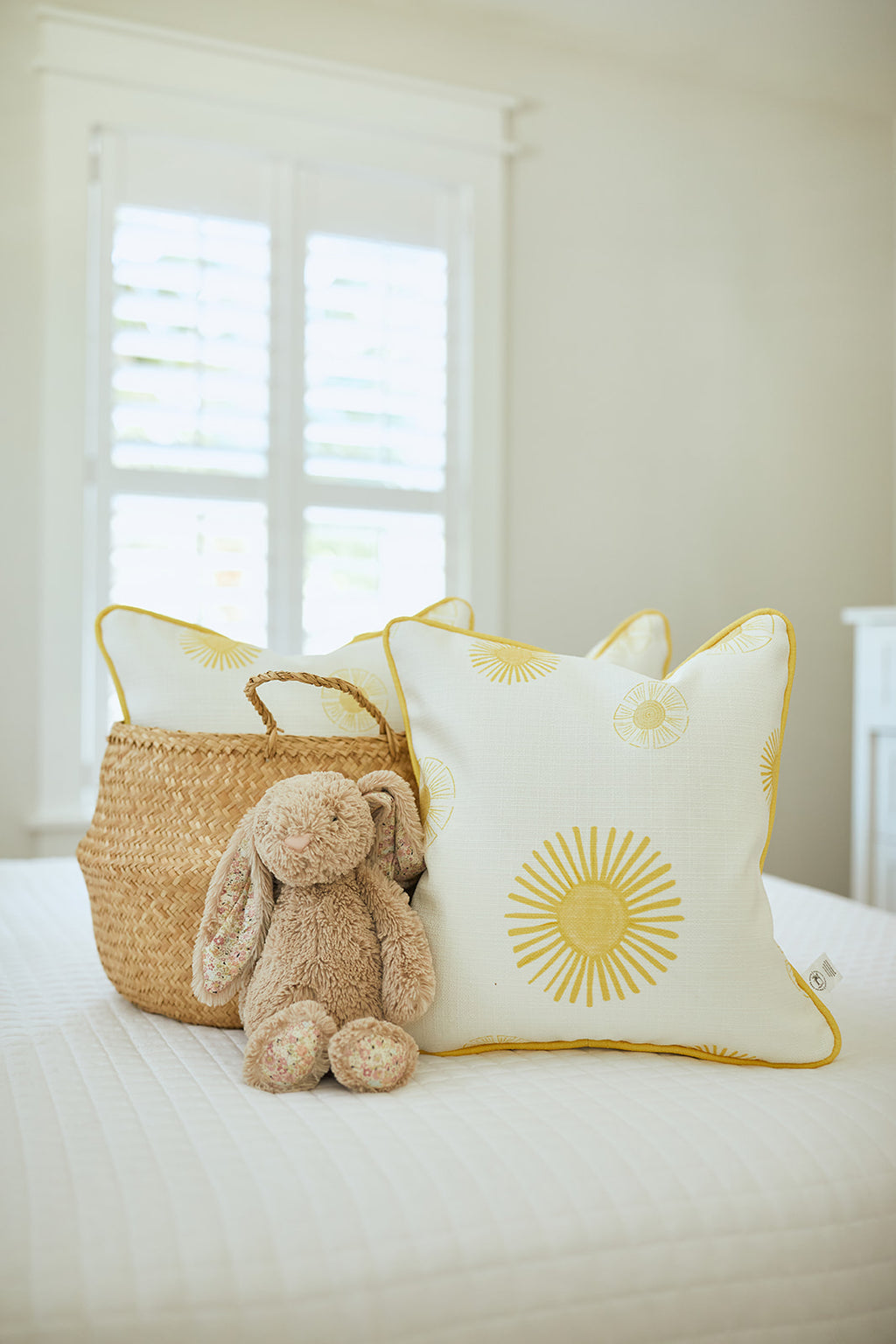 A cozy Kids Collection bedroom scene features the plush Kids Collection: Sun Pillow with sunny patterns, a fluffy bunny, and a woven basket on a white quilted bed. Sunlight streams through white shutters, creating a warm and inviting atmosphere.