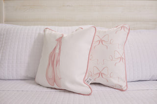 The white quilted bed is adorned with two Kids Collection: Bow & Ballet pillows. One displays ballet shoes, the other pink bows on a light background. Both feature pink piping, standing out against a white wood-paneled wall, highlighting the Bow & Ballet pillow charm.