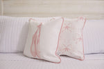 The white quilted bed is adorned with two Kids Collection: Bow & Ballet pillows. One displays ballet shoes, the other pink bows on a light background. Both feature pink piping, standing out against a white wood-paneled wall, highlighting the Bow & Ballet pillow charm.