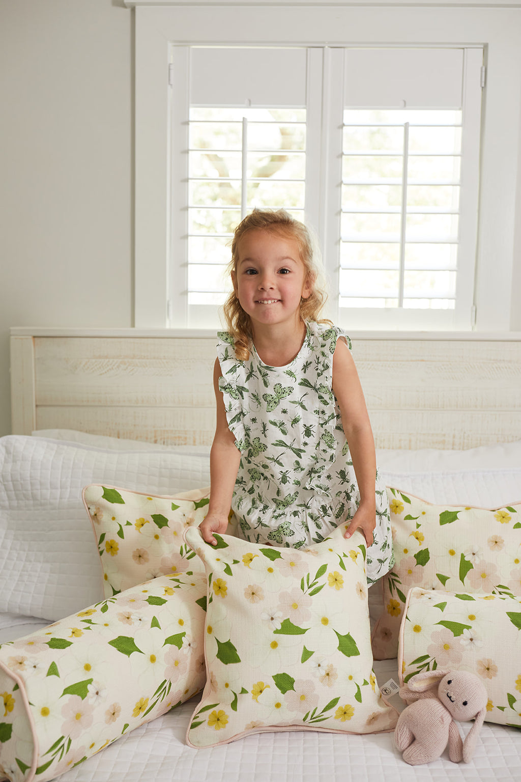 A young blonde girl stands smiling on a bed in a floral dress from the Sewing Down South® Kids Collection, surrounded by pillows, including a Pink Floral Pillow. A small pink teddy bear sits beside her in the bright room with white walls and shuttered windows.