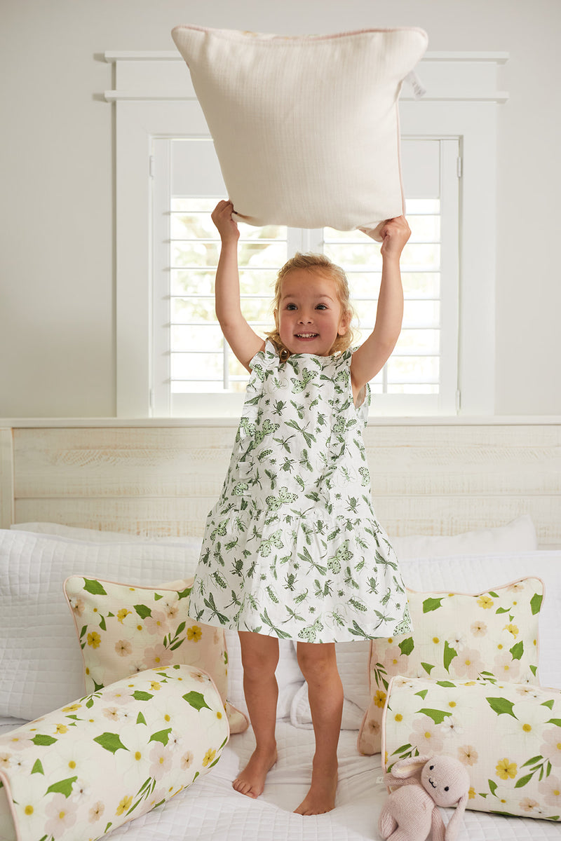 A young girl in a floral dress stands on a bed holding the Kids Collection: Pink Floral Pillow. The bed features matching pillows and a stuffed pink rabbit. Sunlight streams through the window, brightening the cheerful room.