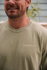 A person wearing the Whats Wrong With My Sewing? Long Sleeve Tee, Sandstone, featuring Sewing Down South, Charleston, South Carolina printed on it. The tees sandstone color complements its soft cotton-like fabric. The blurred background reveals some greenery.