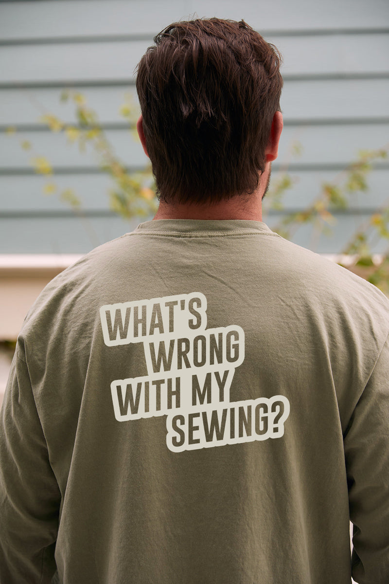 A person with short brown hair is seen from behind, wearing a sandstone long sleeve tee that reads WHATS WRONG WITH MY SEWING? against a light blue wall and some greenery.
