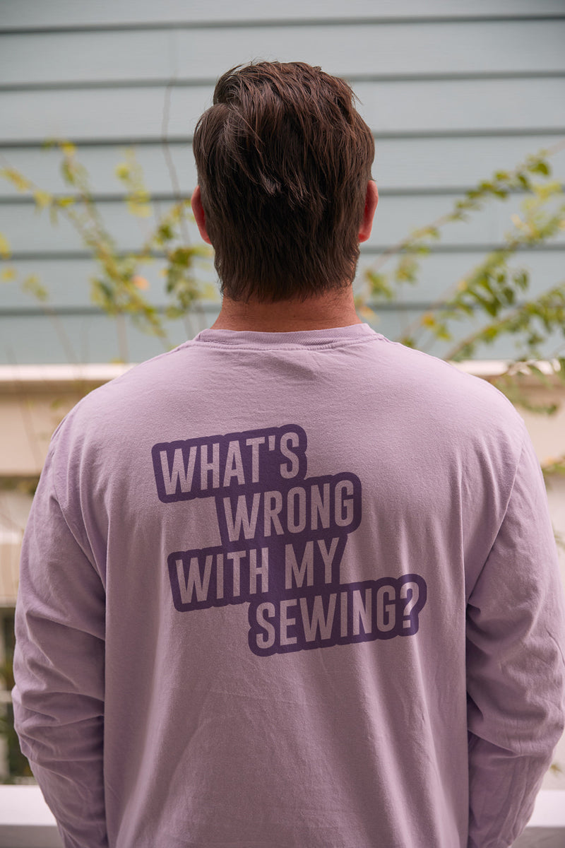 A person with short brown hair, seen from behind, wears the Whats Wrong With My Sewing? Long Sleeve Tee in Orchid. The unisex fit shirt reads the text in dark purple on 100% ring-spun cotton. The backdrop includes a blurred wall and foliage.