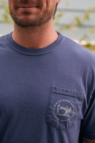 Someone is wearing the SDS Vintage Logo Pocket Tee, a retro-style blue heavyweight cotton shirt. The pocket bears a logo with a sewing machine and Sewing Down South, Charleston, South Carolina 2018 text. The background is lush greenery.