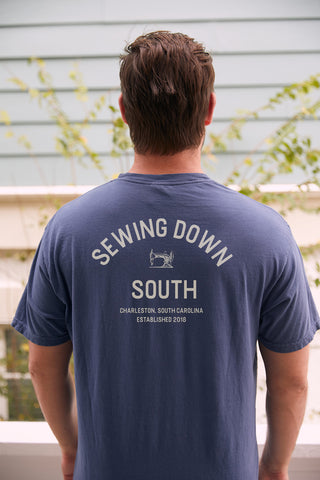 A person, back to the camera, wears an SDS Vintage Logo Pocket Tee in retro blue. It features Sewing Down South, Charleston, South Carolina, Established 2018 and a small sewing machine graphic. Made from heavyweight cotton, they stand against a light building with greenery.