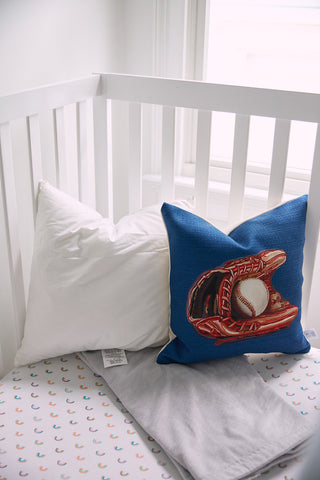 In a cozy crib corner, a vibrant pillow featuring a baseball glove and ball pairs with a plain white pillow and light gray blanket. A patterned crib sheet with small rainbows adds charm. Choose Sewing Down South 17 x 17 Indoor Nursery Inserts for square pillows filled with non-allergenic fiber for safety.