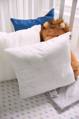 A cozy scene featuring a white crib mattress with a semicircle-patterned sheet, plush brown lion toy, two non-allergenic white pillows, and a Sewing Down South 17 x 17 blue pillow with premium insert, all against white crib bars.