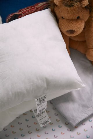 A white pillow with a tag and premium non-allergenic fiber from Sewing Down Souths 17 x 17 Indoor Nursery Inserts rests on a bed with light-patterned sheets. A brown stuffed lion toy is partially visible beside the square nursery pillow.