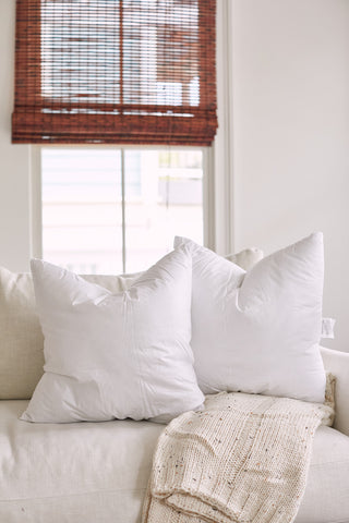 Two Sewing Down South 20 x 20 white pillows filled with non-allergenic fiber rest on a beige sofa next to a knitted beige blanket. Bamboo blinds softly filter the light through the window, creating a cozy and inviting atmosphere.