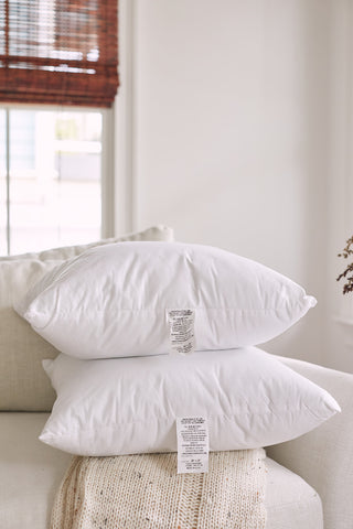 Two Sewing Down South 20 x 20 Indoor Inserts are stacked on a beige sofa, with a knitted blanket underneath. Tags with care instructions hang from the pillows, featuring non-allergenic fibers. Brown-blinded windows in the background let natural light illuminate the cozy scene.