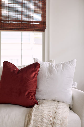 The cozy living room showcases a beige sofa with a red velvet pillow and a white pillow, each filled with Sewing Down South 20 x 20 Indoor Inserts. A knitted beige throw graces the armrest, while bamboo blinds softly diffuse natural light through the window.