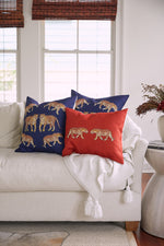 A white sofa features a white tassel throw with three exotic pillows: two blue On The Prowl Midnight animal-patterned and one orange. A nearby side table with dried flowers is framed by bright windows in the background.