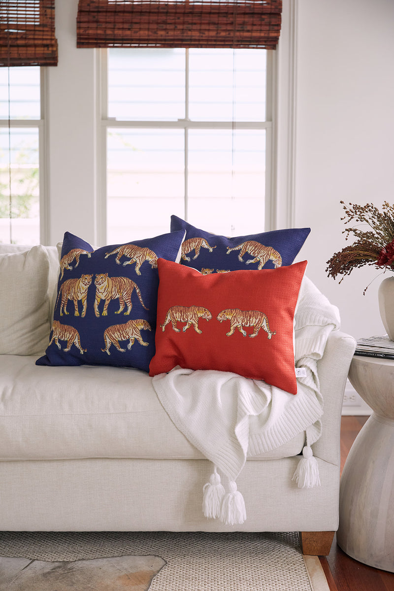 A white sofa features a white tassel throw with three exotic pillows: two blue On The Prowl Midnight animal-patterned and one orange. A nearby side table with dried flowers is framed by bright windows in the background.