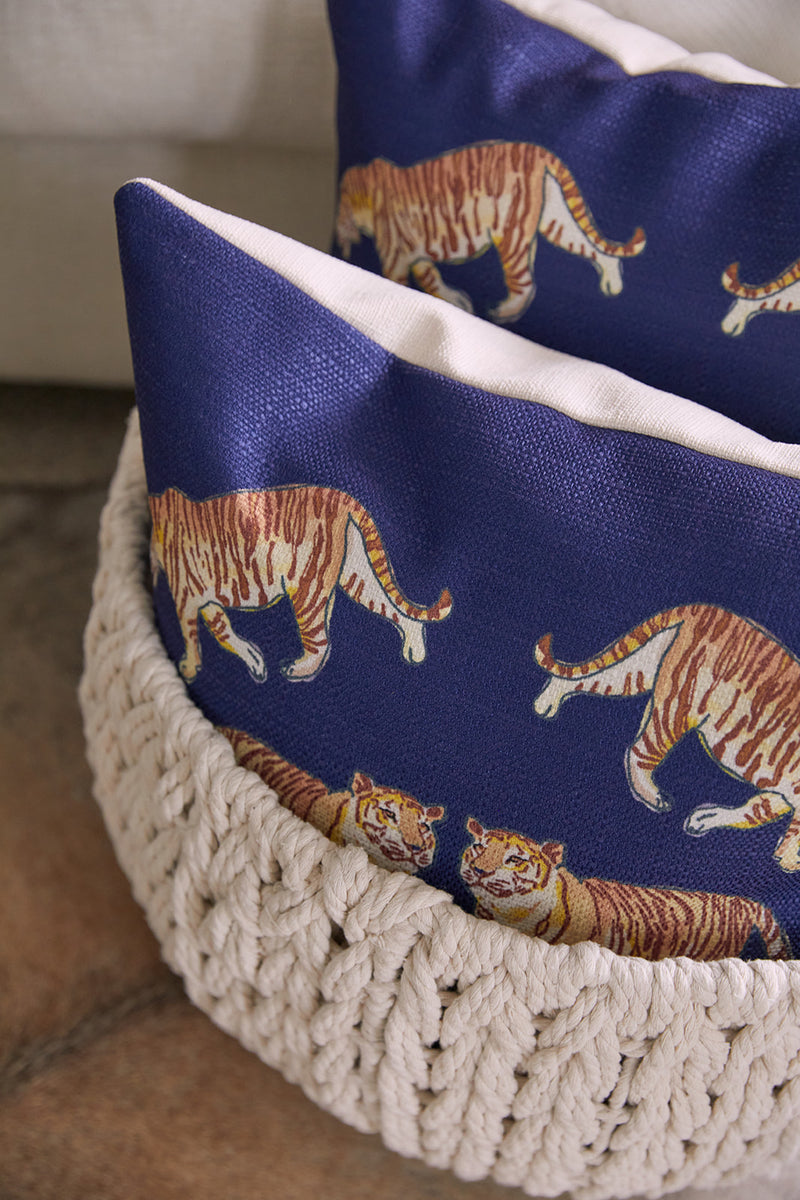 Two On The Prowl Midnight throw pillows with orange tiger patterns sit in a cream woven basket on a soft brown textile, adding an exotic flair.