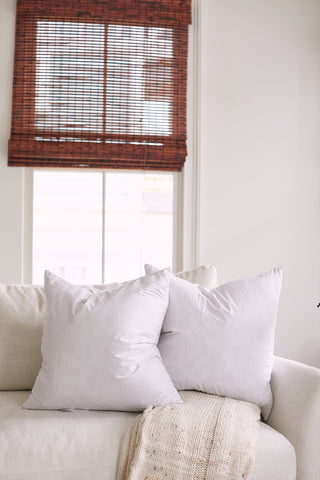 A beige sofa with two large, white Sewing Down South 20 x 20 Indoor Inserts rests in front of a window with bamboo blinds. A beige throw drapes over the armrest, and soft light filters through the window.
