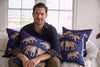 A person sits on a couch, surrounded by exotic On The Prowl Midnight Pillows with tiger designs. The room is brightly lit by a large window. They wear a dark shirt and smile slightly at the camera.