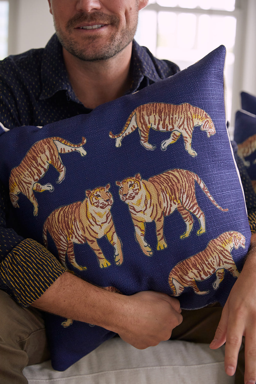 A person in a dark shirt holds the On The Prowl Midnight Pillow, decorated with tiger illustrations for an exotic touch. They sit against a softly lit backdrop, highlighting the animal pattern.
