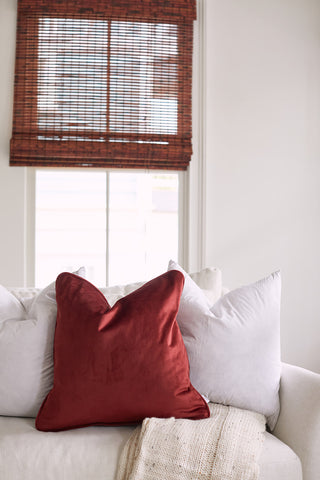 A white couch with a red cushion and two white cushions, featuring Sewing Down South 20 x 20 Indoor Inserts, sits before a window with brown bamboo blinds. A beige knitted throw drapes over the side, amid bright, neutral walls and surroundings.