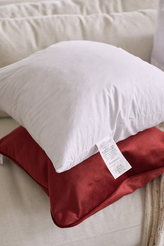 A white Sewing Down South 20 x 20 pillow with a non-allergenic fiber insert rests on a red pillow, both cozily set on a light-colored sofa.