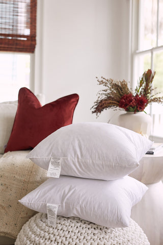 Two Sewing Down South 20 x 20 Indoor Inserts, featuring non-allergenic fiber, are stacked on a textured white knit pouf in a cozy living space. A red cushion and a neutral knit blanket are on the sofa. A vase with red flowers and dried grasses decorates a round white table. Soft natural light fills the room.