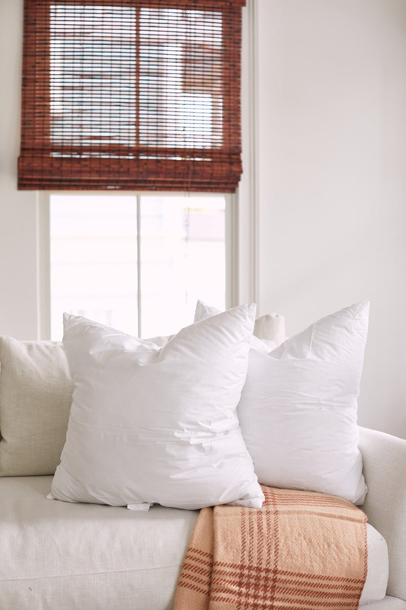 A cozy white sofa with Sewing Down South 23 x 23 Indoor Inserts nestled in two large pillows and a beige plaid throw blanket draped neatly on it. Brown bamboo blinds on the window filter soft light into the room, creating a serene atmosphere.