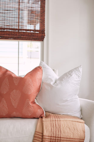 A cozy living room scene showcases a white sofa with two pillows: one large using a Sewing Down South 23 x 23 Indoor Insert and a smaller orange pillow with a leaf pattern. Nearby, a plaid blanket is draped, and bamboo shades on the window are partially pulled down.