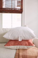 A white pillow with a Sewing Down South 23 x 23 Indoor Insert is stacked on a peach chevron-patterned throw pillow. Both sit on a beige sofa with a plaid blanket, framed by wooden-blinded windows that enhance the cozy atmosphere.