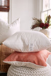 A cozy scene features two pillows stacked: a Sewing Down South 23 x 23 Indoor Insert in white atop an orange one with a white leaf pattern. A cream pillow, dried flowers by the window, and an oversized square insert knitted pouf complete the look.