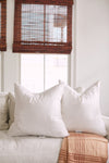 A cozy living room highlights a white sofa with two large pillows featuring Sewing Down South 23 x 23 Indoor Inserts. A plaid blanket in brown and cream drapes over the sofa, while wooden-blind windows softly filter light in the background.