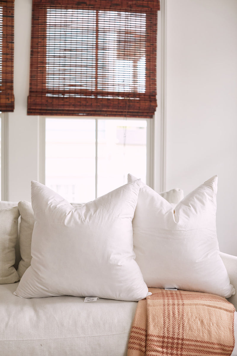 A cozy living room highlights a white sofa with two large pillows featuring Sewing Down South 23 x 23 Indoor Inserts. A plaid blanket in brown and cream drapes over the sofa, while wooden-blind windows softly filter light in the background.