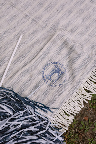 Close-up of a textured, oversized SDS Embroidered Picnic Blanket with Tassels featuring a blue bell emblem. A pom-pom and white stick lie next to grass. The blanket showcases a striped, woven pattern.