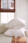 Two white pillows with Sewing Down South 23 x 23 Indoor Inserts are stacked on a light couch in a bright room with natural light. A window with wooden blinds and a checked throw blanket draped over the couch arm provide the backdrop, and tags are attached to the pillows.