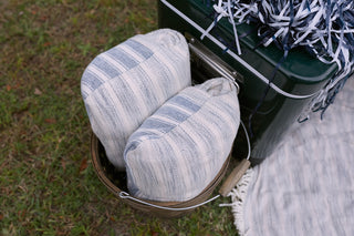 A metal pail holds two yarn-dyed stripe pattern blankets in white and blue, nestled on grass beside a green container, partly hidden by blue and white pom-poms and an elevated SDS Embroidered Picnic Blanket with Tassels.