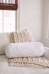 A cozy armchair with a cream knit blanket and a white cylindrical pillow featuring the Sewing Down South 8 x 20 Bolster Indoor Insert sits near a window with natural light and bamboo blinds.