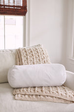 A cozy armchair with a cream knit blanket and a white cylindrical pillow featuring the Sewing Down South 8 x 20 Bolster Indoor Insert sits near a window with natural light and bamboo blinds.