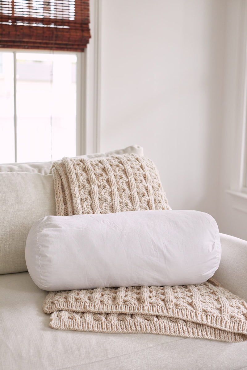 A cozy armchair with a cream knit blanket and a white cylindrical pillow featuring the Sewing Down South 8 x 20 Bolster Indoor Insert sits near a window with natural light and bamboo blinds.