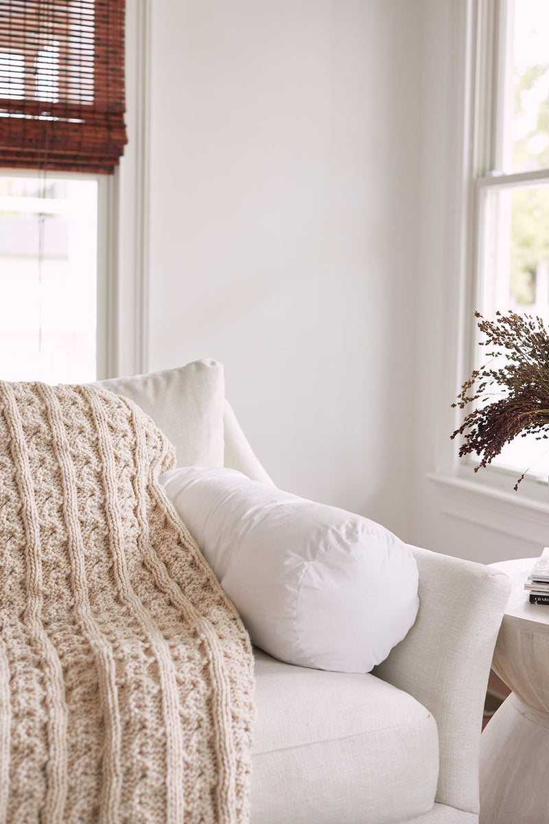 A cozy cream armchair with a textured beige knit blanket and a white bolster pillow with a Sewing Down South 8 x 20 Bolster Indoor Insert sits by a window with wooden blinds and dried flowers in a vase on the side table.