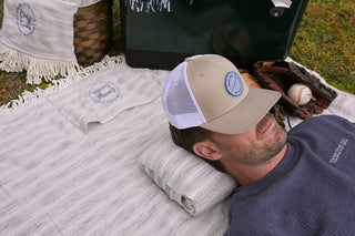 A man wearing a cap relaxes on an oversized SDS Embroidered Picnic Blanket with Tassels, its yarn-dyed stripe pattern adding vibrancy. A rolled-up towel acts as his pillow. Nearby, a baseball glove with a ball, a woven basket, and a cooler complete the scene. Text on his cap and shirt hint at the days adventure.