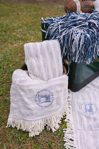 A picnic scene with SDS Embroidered Picnic Blankets with Tassels in white and gray stripes, one folded in a green bucket. A pom-pom and vintage football sit nearby on the grass. These blankets, featuring a horse logo and fringed edges, enhance the picnic experience.