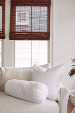 A white couch with a large pillow filled with a non-allergenic fiber insert and a Sewing Down South 8 x 20 Bolster Indoor Insert sits in front of two windows with brown bamboo blinds in a bright, airy room.
