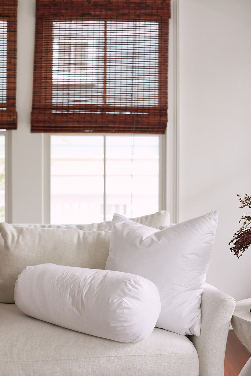 A white couch with a large pillow filled with a non-allergenic fiber insert and a Sewing Down South 8 x 20 Bolster Indoor Insert sits in front of two windows with brown bamboo blinds in a bright, airy room.