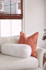 A white sofa features the Sewing Down South 8 x 20 Bolster Indoor Insert and a coral leaf-pattern throw pillow. Sunlight streams through wooden blinds, highlighting the non-allergenic fiber fill of the bolster insert.