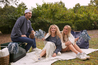 Three friends enjoy a picnic, with one man seated on a cooler holding a football and two women waving pom-poms on an SDS Embroidered Picnic Blanket with Tassels. They are smiling and casually dressed, surrounded by lush greenery.