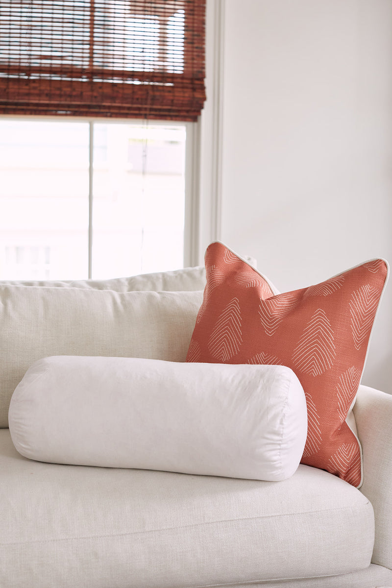 A Sewing Down South 8 x 20 Bolster Indoor Insert and an orange pillow with a subtle leaf pattern sit on a light-colored sofa near a window with bamboo blinds.